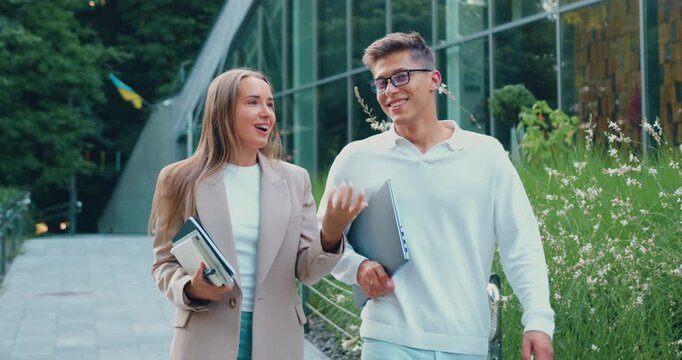 Young professionals walking and talking outside modern campus building with laptop and notebooks, smiling and gesturing in daylight, green landscaping, casual business attire, friendly conversation