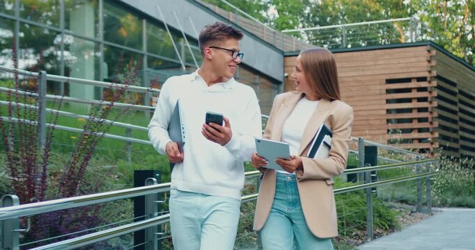 Young professionals man and woman walking outdoors sharing smartphone and tablet, laughing and discussing on campus walkway with modern buildings and greenery. Soft natural light highlights scene