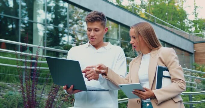Young business people collaborating outdoors with laptop and notebook near modern glass building, smiling and pointing at screen while discussing project details, daylight urban campus setting