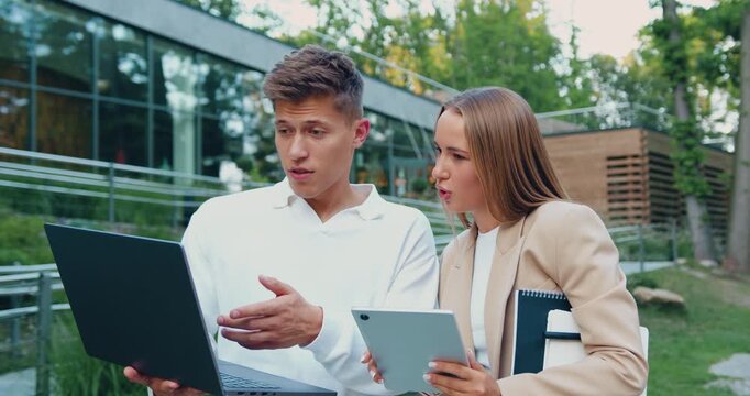 Man and woman students studying together, using laptop, makes of distance learning walking by university campus. Male and female freelancer working on outside during break time