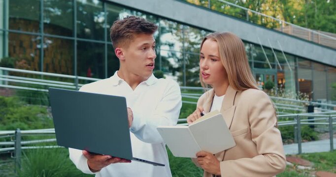 Close-up of a businessman and businesswoman in formal attire standing outside a modern office building, discussing work-related topics using a digital tablet and handwritten notes in a notebook