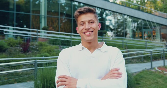 Young man outdoors with arms crossed smiling and shifting expression in front of modern glass building, daytime portrait motion scene confident casual pose, urban park setting, natural sunlight