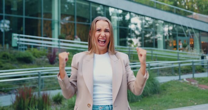 Young woman celebrating with raised fists and joyful smile in front of modern glass building, outdoor daylight midshot, confident upbeat mood, visible movement and expression of victory