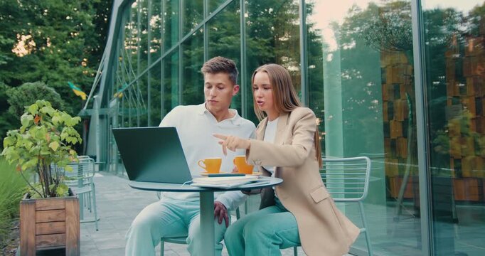 Students man and woman working together on a laptop, discussing a project in an outdoor workspace near a contemporary university campus.