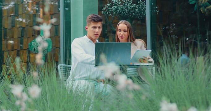 Two friends man and woman reacting with surprise and laughter while viewing open laptop at outdoor cafe in morning light, leafy foreground blur and glass storefront background, candid social