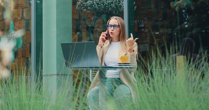 Young Caucasian businesswoman in casual clothes talking on mobile phone outdoors while sitting on cafe near office building. Online chat.