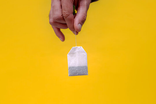 Hand holding a tea bag. A white paper tea bag hangs by its string against a vivid yellow background. Suits food, beverage, wellness, and packaging projects.