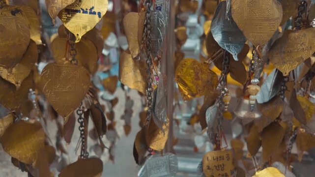 golden bodhi leaf wishing ornaments hanging on chains at temple, silver metal prayer tags engraved with handwritten messages at buddhist shrine 4k