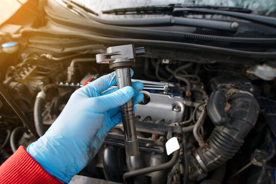 Gloved hand holding car ignition coil in engine bay, mechanic inspecting ignition coil during auto repair service. Close up of car ignition coil, automotive ignition system component replacement