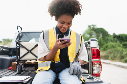 Female field worker in a high visibility vest checks a smartphone while sitting in a utility truck bed, showing mobile coordination, outdoor operations, workplace safety, and field service.