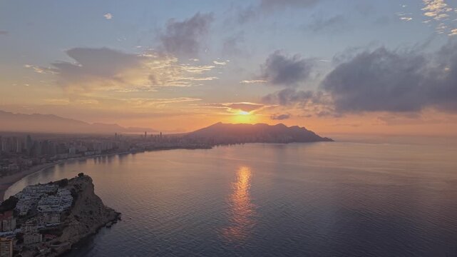 Aerial view of the Benidorm coastline at sunset with the sun setting behind mountains, reflecting on the sea and illuminating the city in Benidorm, Valencian Community, Spain.