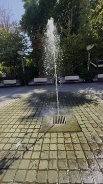 Refreshing water fountain in a sunny urban park with cobblestone ground and green trees in