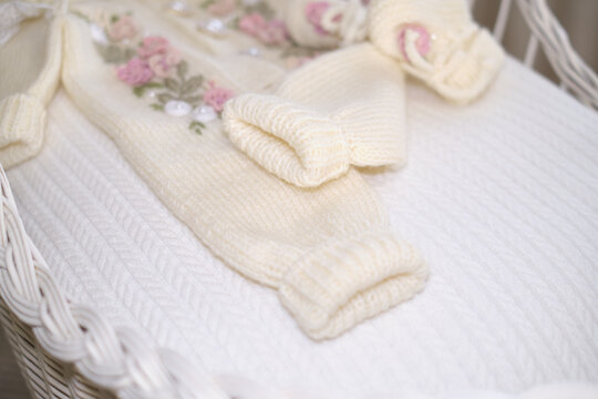 A cream knitted baby outfit lies in a white wicker cradle, showing soft cuffs and floral details. The image reflects newborn preparation, handmade care, family tenderness, and gentle anticipation.