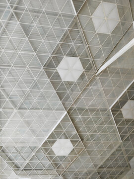 Repeated triangular ceiling members surround pale polygonal panels, creating a delicate overhead pattern within a spacious building