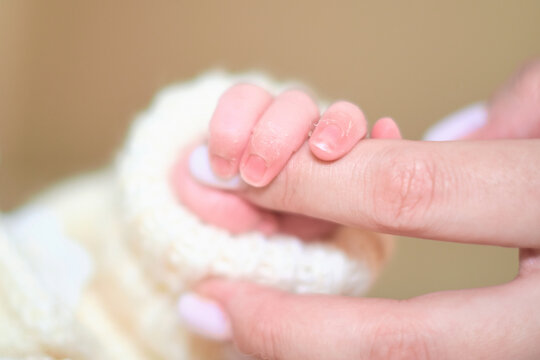 A newborn baby gently grips an adult finger with tiny curled fingers, resting against cream knitwear in soft focus. The image reflects trust, early bonding, parental care, and emotional safety.