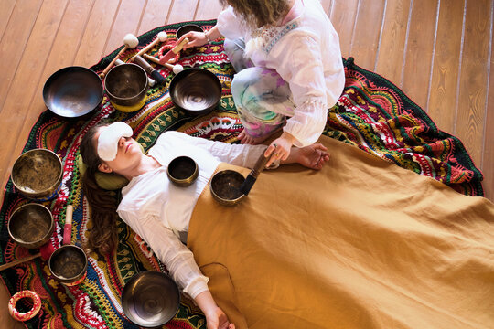 From above, a client lies under a blanket during a restorative sound bath while a therapist works with bowls and soft mallets. The image connects to burnout recovery, relaxation, and wellness trends.