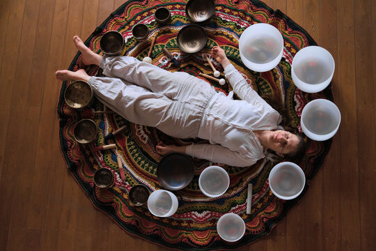Top down view of a sound bath session with a woman lying on a colorful rug among crystal bowls, Tibetan bowls, and mallets. Vibrational rituals support rest, clarity, and stress relief.