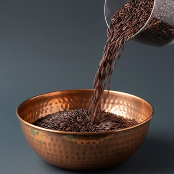 Raw black rice pouring from clear glass cup into hammered copper bowl on dark background