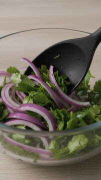 Black spoon mixing sliced red onion and fresh cilantro salad in clear glass bowl
