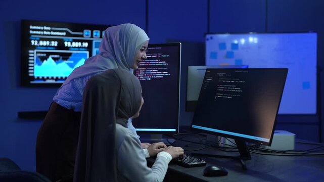 Two Muslim female software developers in hijab coding on monitors in dark office, Happy programmer team working on IT system, Modern technology, teamwork, cyber security and data development concepts