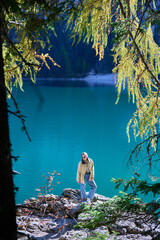 Young woman poses on lakeside rocks beneath sunlit larch canopy overlooking turquoise Lago di Braies. Vertical composition suits travel campaigns, wellness themes and autumn destination marketing © anatoliy_gleb