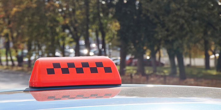 Taxi roof sign with black checker pattern marking urban transportation above a parked car, reflecting daylight beneath leafy street trees