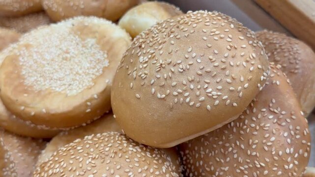 Fresh sesame burger buns lie on baking paper inside bakery shop display. Golden baked bread rolls create fast food ingredient showcase, grocery retail assortment and culinary preparation product.