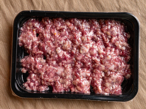 fresh raw minced pork in a black plastic tray wrapped in film on a beige woven fabric surface, uncooked ground meat with white fat ready for cooking, food ingredient