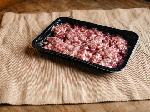fresh raw minced pork in a black plastic tray wrapped in film on a beige woven fabric surface, uncooked ground meat with white fat ready for cooking, food ingredient