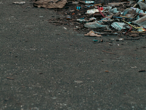 Pile of garbage and litter on the edge of a dark asphalt road, scattered plastic wrappers, dry leaves and wood debris, urban pollution scene with empty copy space on the left