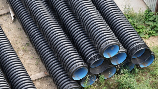 Stacks of black corrugated plastic drainage pipes with blue inner walls stored outdoors, showing construction materials prepared for underground drainage and utility infrastructure projects