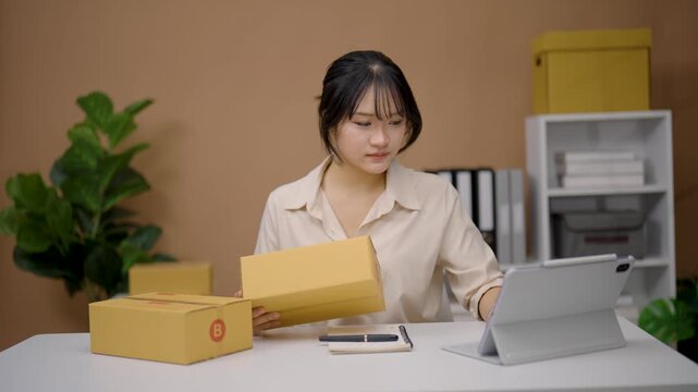 Young woman working at desk with cardboard box holding device tablet organizing parcel for shipping or home business packing