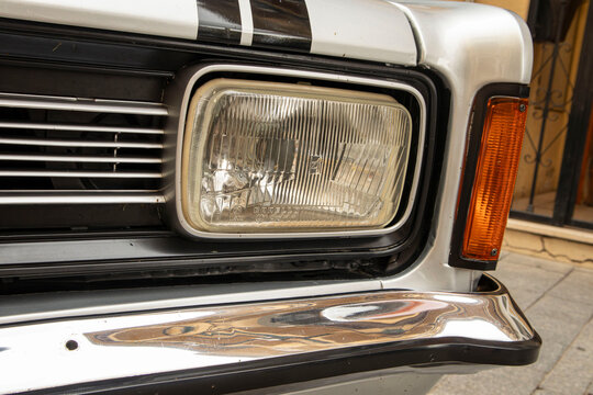 GANDIA, SPAIN - OCTOBER 4, 2026: Rectangular Carello headlight, amber turn signal, and chrome bumper detail on a vintage silver classic car.