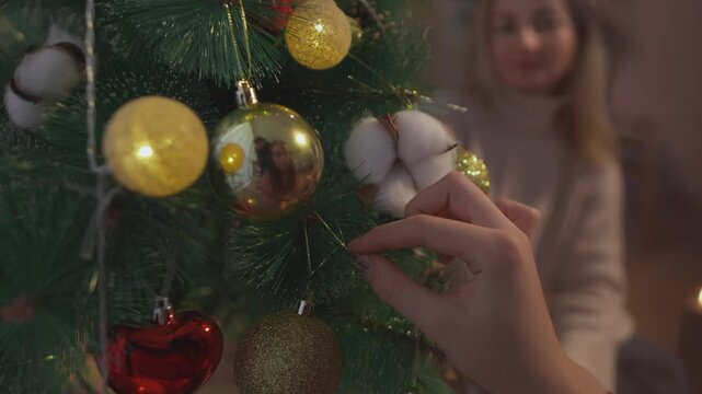 Detailed holiday footage captures hands as they hang shiny gold baubles among green branches and warm fairy lights. Soft bokeh view presents traditional tree decorating with strong commercial appeal.