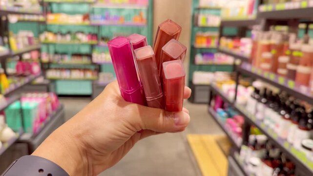 Hand Holding Several Colorful Lipsticks in Cosmetics Store &ndash; Choosing Between Different Shades Against Makeup Shelves