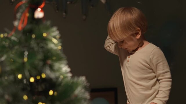 At Christmas, a toddler boy explores festive objects beside the Christmas tree. The medium side view highlights curious toddler, seasonal discovery and seasonal joy for lifestyle media.