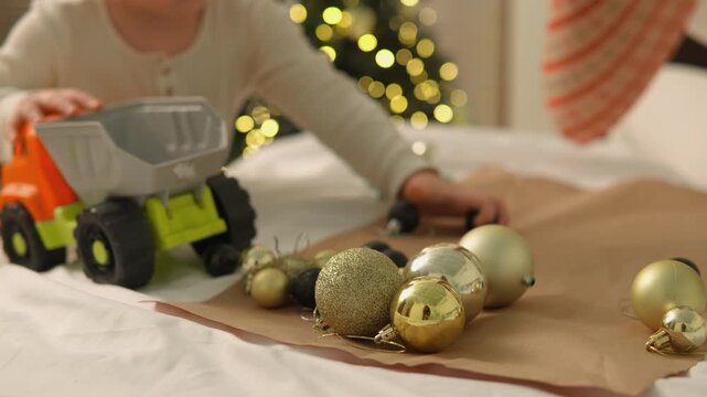 A toddler boy plays with a toy truck and ornaments while his mother joins him in a festive home. The wide floor view conveys shared play, responsive parenting and family warmth.