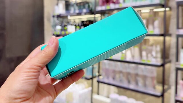 Hand Holding Shampoo Bottle and Hair Care Box in Cosmetics Store &ndash; Hair Oil, Tinted Balm and Conditioner Against Hair Care Shelves