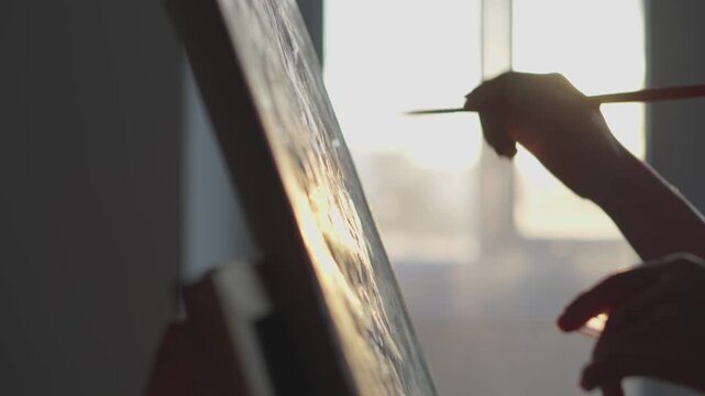 Close up of an artist's hand holding a brush while painting on a canvas, illuminated by soft sunlight. The scene highlights creativity, inspiration, and the beauty of artistic work