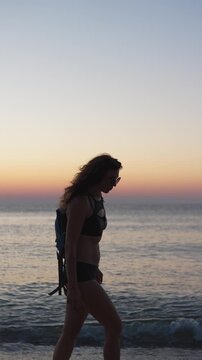 A woman in swimwear strolls on the shoreline with gentle waves and a colorful sunset sky in the background. The serene atmosphere highlights relaxation and summer travel vibes