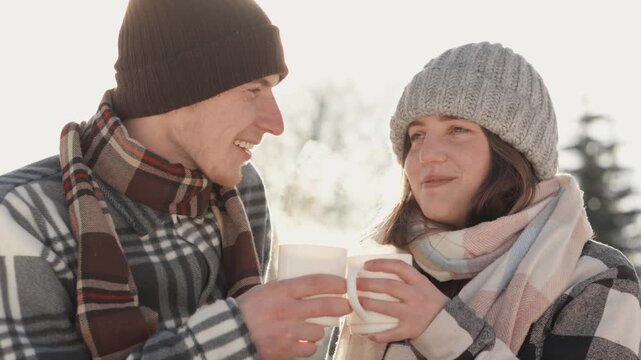 A cheerful couple dressed in hats and scarves clink mugs while standing outside in bright winter sunlight. Their laughter and connection capture the warmth and romance of winter togetherness
