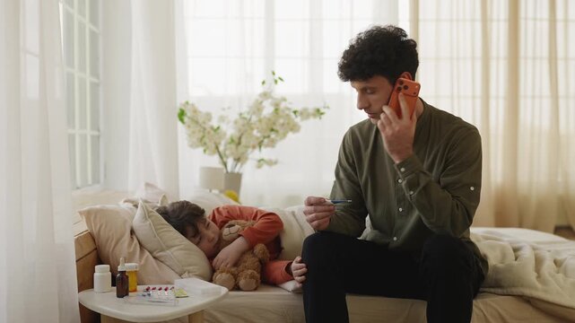 Worried dad calls doctor and checks thermometer while sick son lies in bed