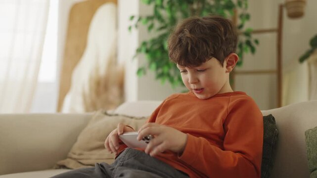 Cheerful little boy plays video game on phone while resting on sofa at home