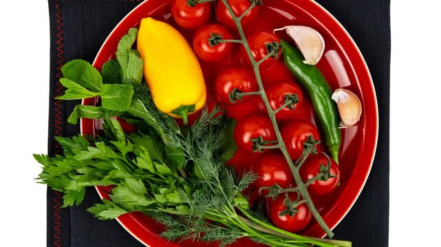 Bright, fresh vegetables and herbs arranged neatly on a red plate, including vibrant yellow bell pepper, ripe red cherry tomatoes on the vine, green chili pepper, garlic cloves, and an assortment of.