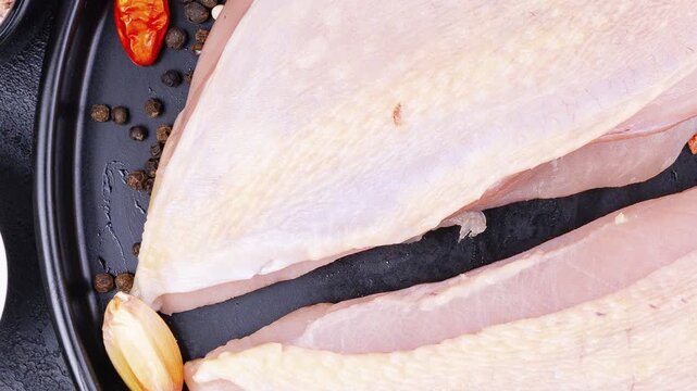 Raw turkey meat placed on a black plate, accompanied by a small bowl of mixed peppercorns and garlic cloves.