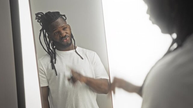Young man points finger at his mirror reflection while looking into the glass, wearing a white tshirt in a studio; self reflection confidence.