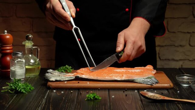 Cook uses culinary fork and a knife to slice a fresh salmon fillet on a wooden cutting board, surrounded by herbs, oil, seasonings, and utensils in a rustic kitchen