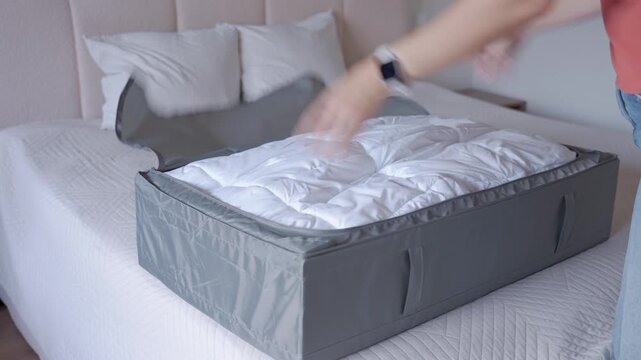Person opening gray fabric storage bag and taking out white duvet on bed. Organizing bedding and bedroom storage