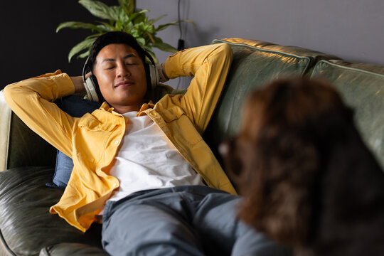 Mid adult Asian man reclining on green leather sofa at home wearing headphones, blurred dog nearby