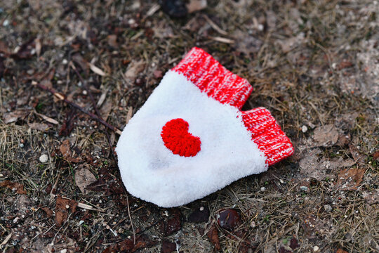 White knitted mitten with red embroidered heart and ribbed cuff.  Solitary mitten with bright red heart lies forgotten on dry earth, symbolizing enduring love abandoned in harsh reality of cold world.